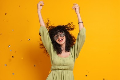 Happy young woman wearing carnival mask and confetti on yellow background