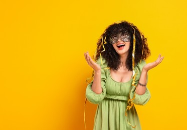 Happy young woman wearing carnival mask and tinsel on yellow background, space for text