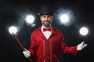 Photo of Portrait of showman in red costume, cane and hat on black background with spotlights and smoke