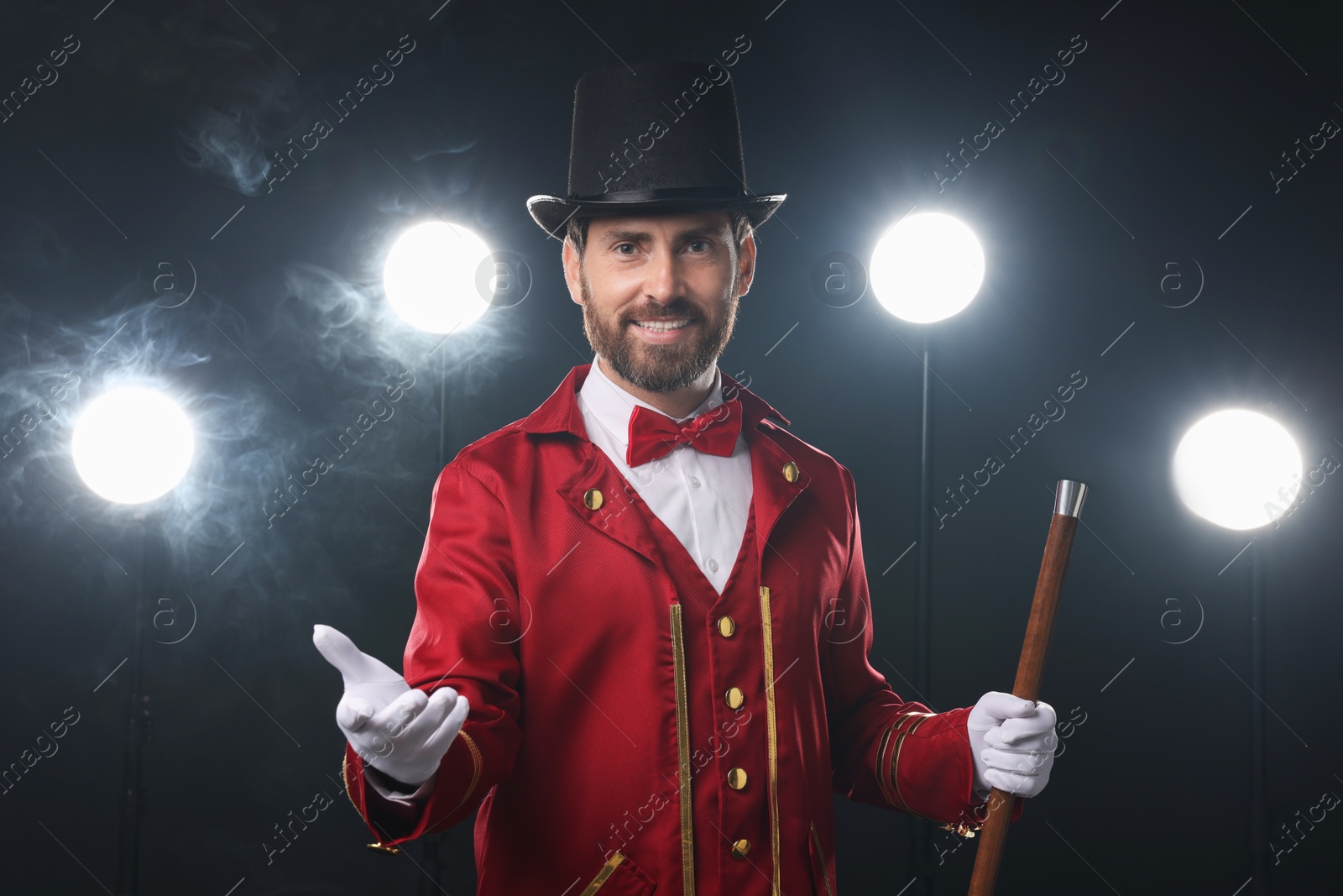 Photo of Portrait of showman in red costume, cane and hat on black background with spotlights and smoke