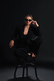 Beautiful woman in stylish suit sitting on chair against black background