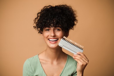 Happy young woman with teeth color samples on beige background. Dental veneers