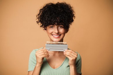 Happy young woman with teeth color samples on beige background. Dental veneers