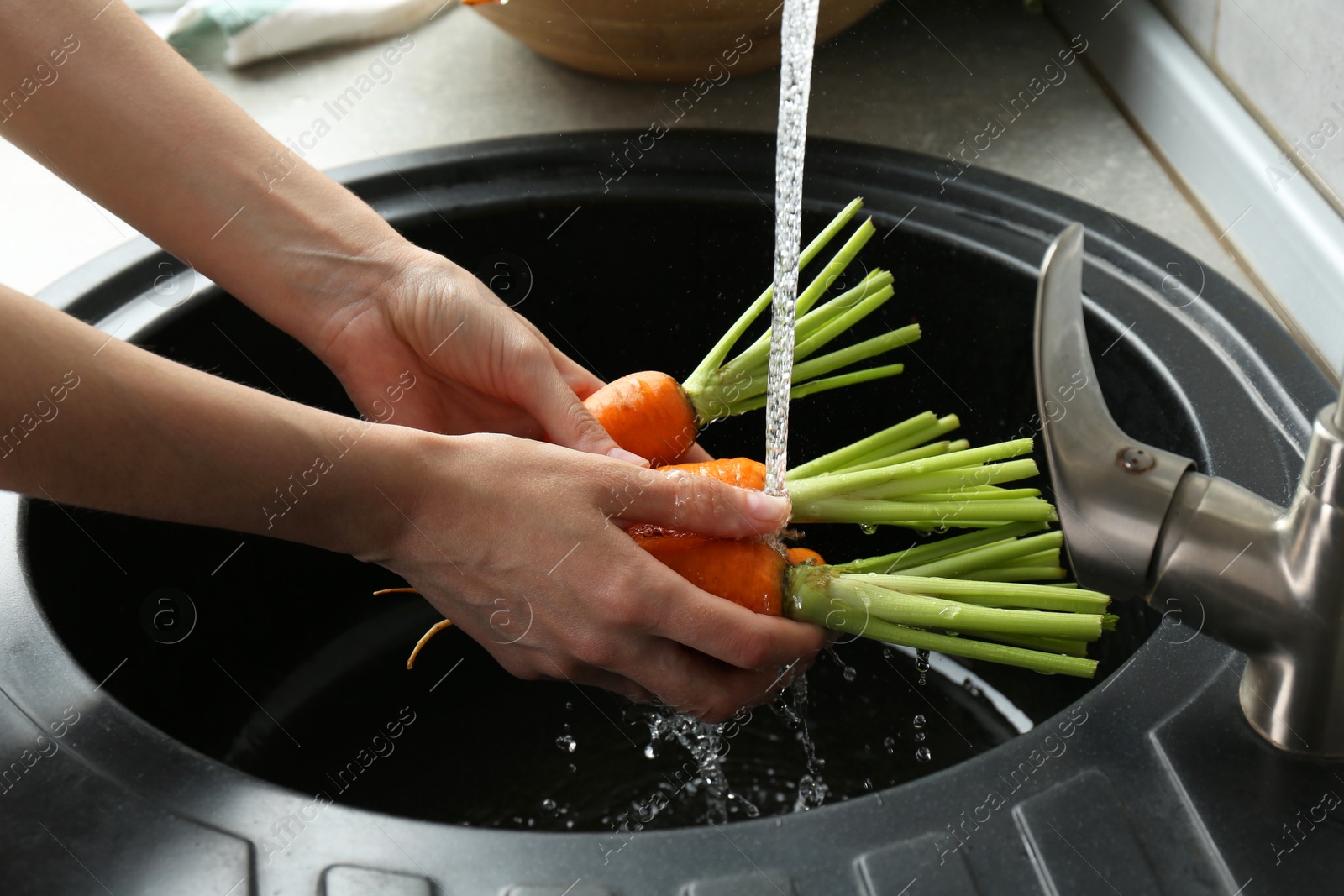 Photo of Woman washing fresh carrots under tap water in above sink indoors, closeup