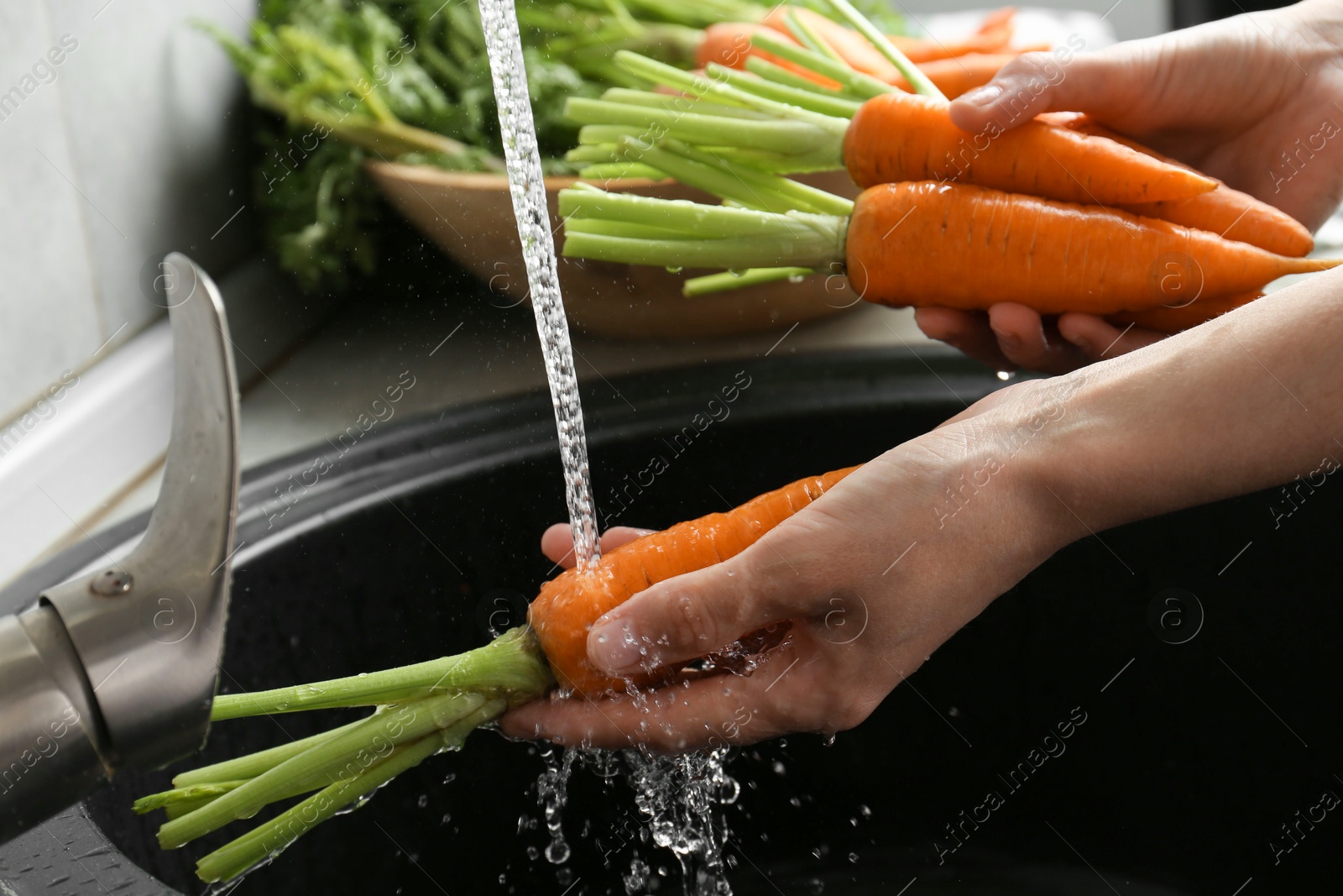 Photo of Woman washing fresh carrots under tap water in above sink indoors, closeup