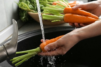 Photo of Woman washing fresh carrots under tap water in above sink indoors, closeup