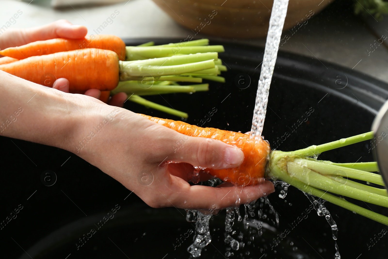 Photo of Woman washing fresh carrots under tap water in above sink indoors, closeup