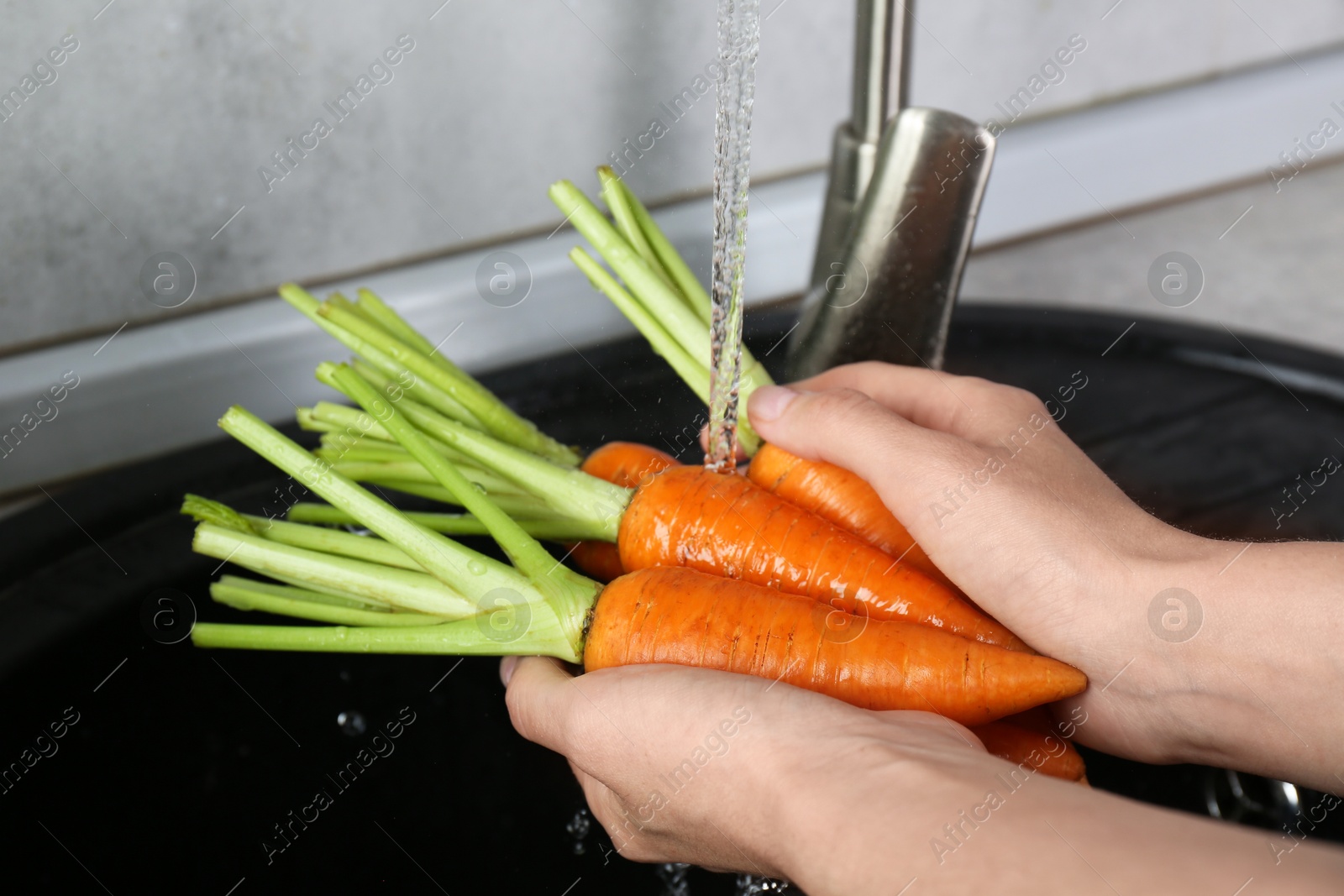 Photo of Woman washing fresh carrots under tap water in above sink indoors, closeup