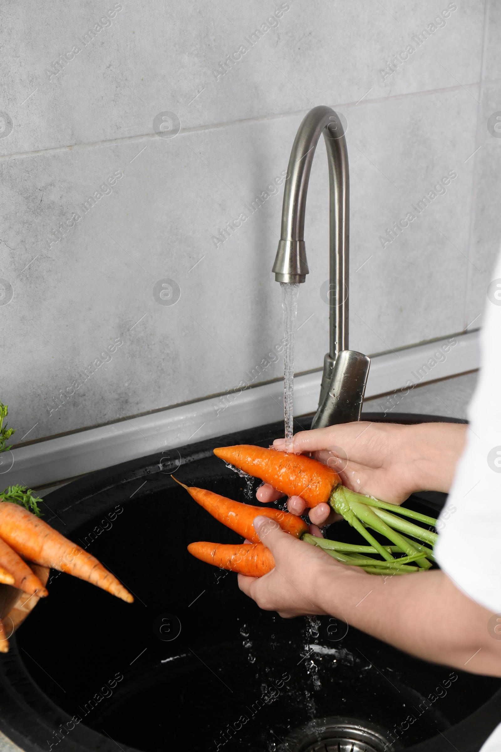 Photo of Woman washing fresh carrots under tap water in above sink indoors, closeup