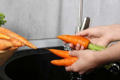 Photo of Woman washing fresh carrots under tap water in above sink indoors, closeup