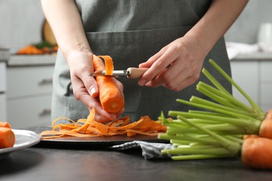 Woman peeling fresh carrot at dark gray table in kitchen, closeup