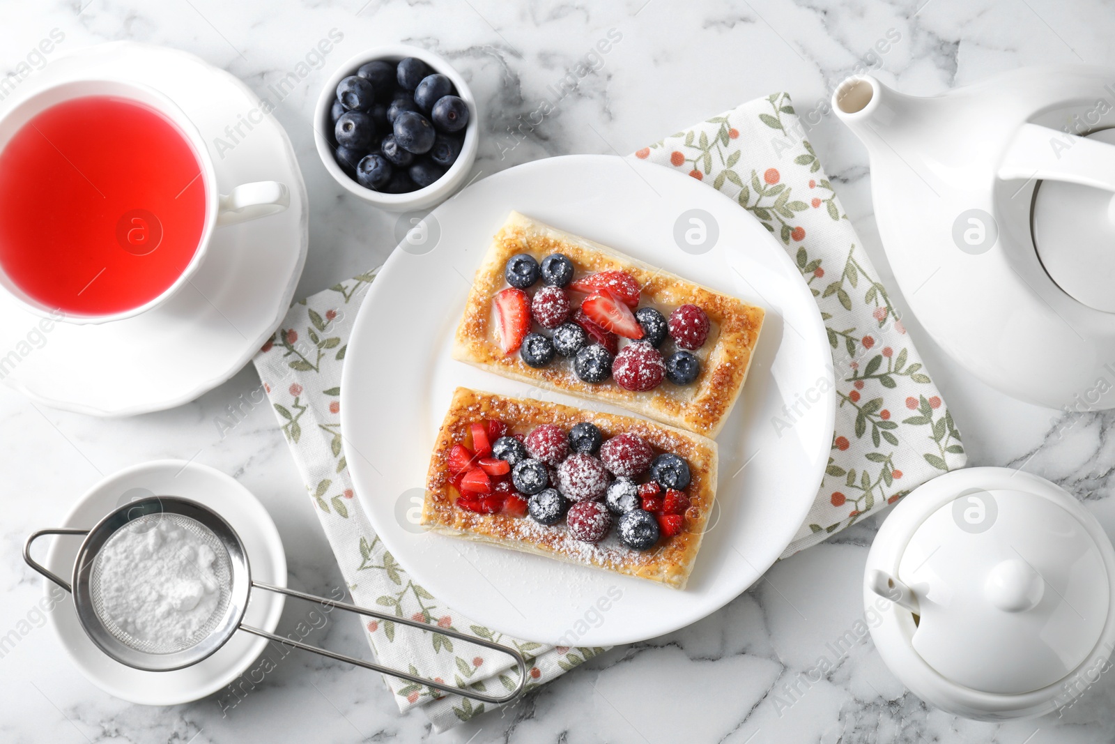 Photo of Tasty puff pastries with berries and tea on white marble table, flat lay