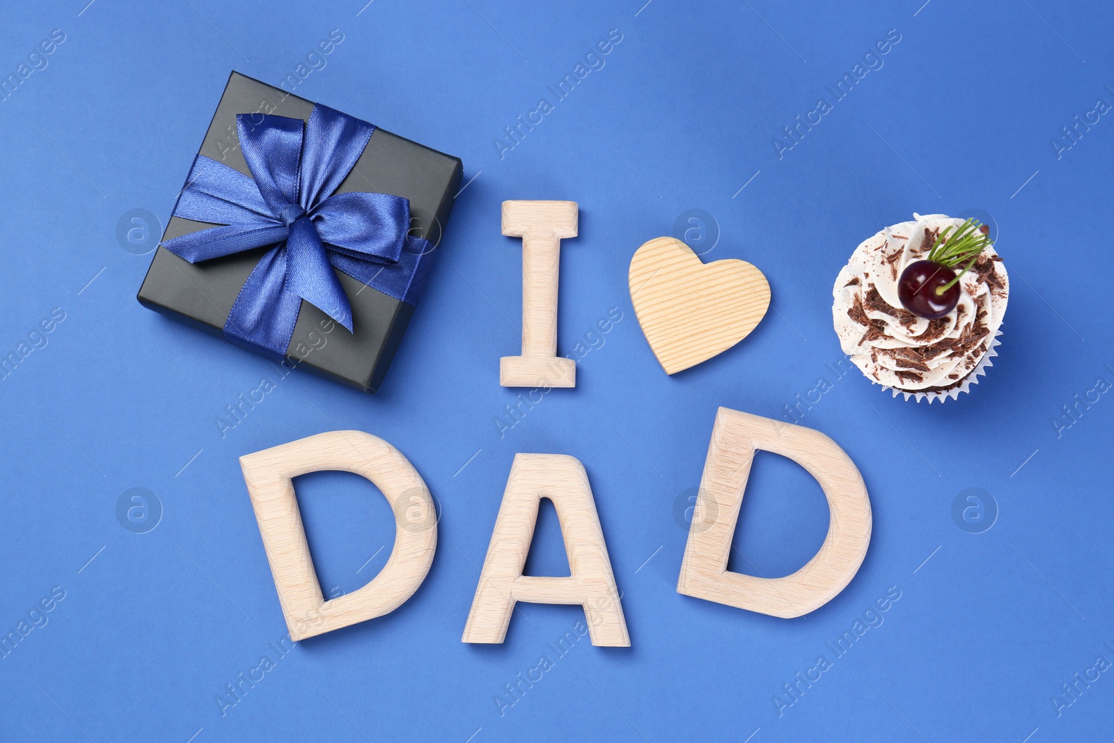 Photo of Happy Father's Day. Phrase I love Dad made with wooden letters, decorative heart, gift box and cupcake on blue background, flat lay