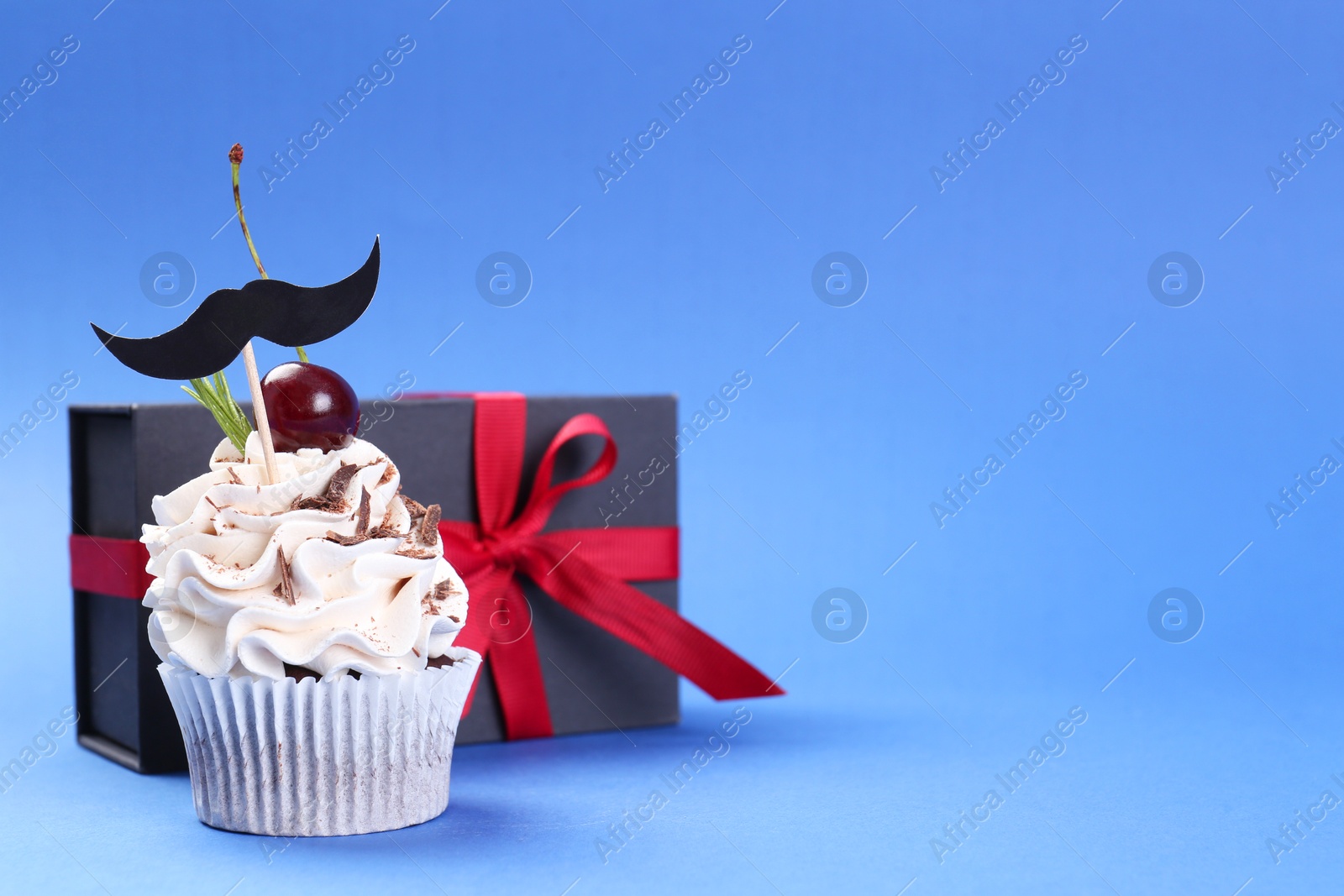 Photo of Happy Father's Day. Cupcake with mustache topper and gift box on blue background. Space for text