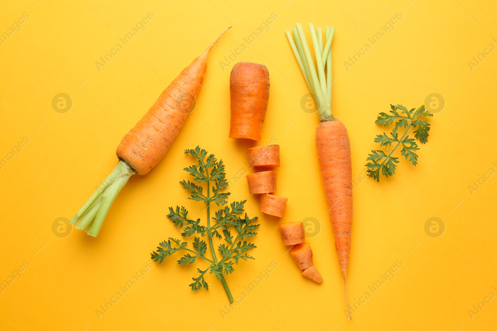 Photo of Whole and cut fresh carrots with green leaves on yellow background, flat lay