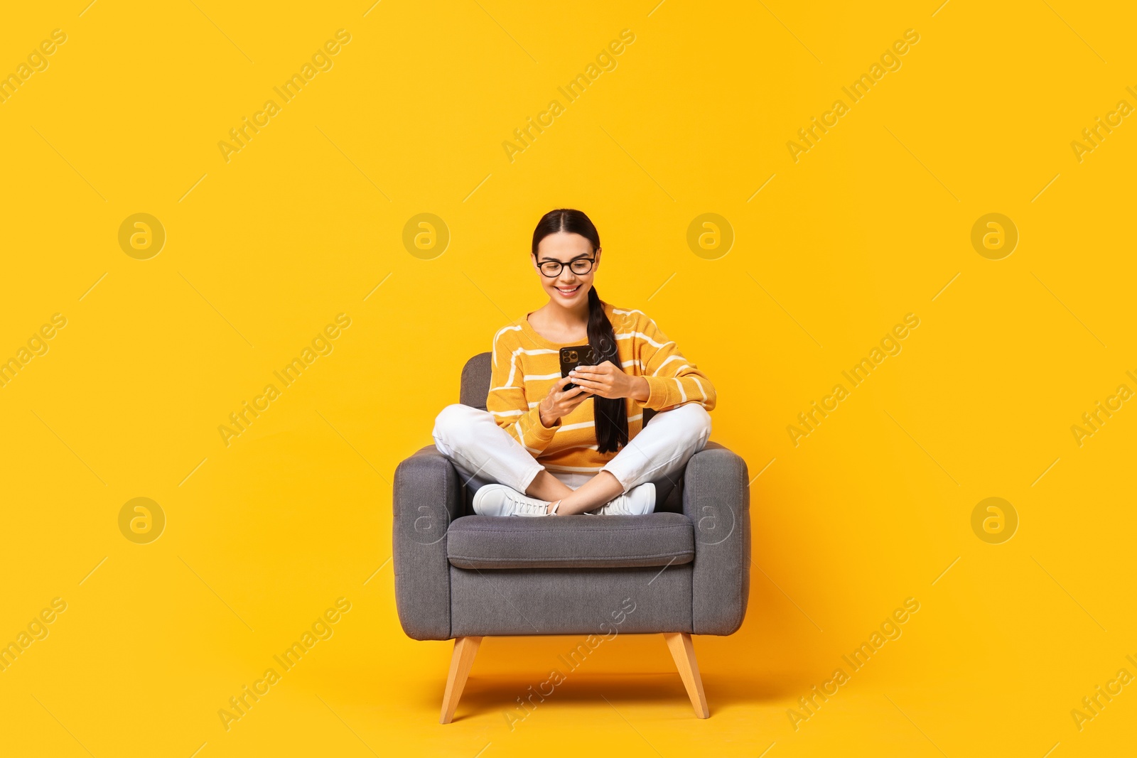 Photo of Smiling woman with smartphone sitting on armchair against yellow background