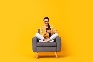 Photo of Smiling woman with smartphone sitting on armchair against yellow background