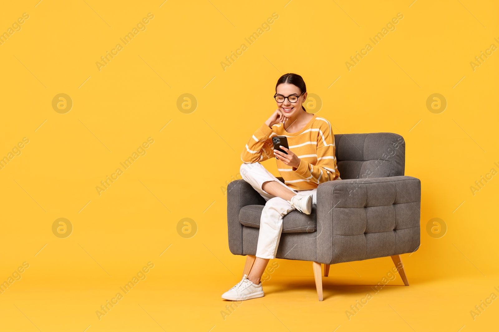 Photo of Smiling woman with smartphone sitting on armchair against yellow background. Space for text