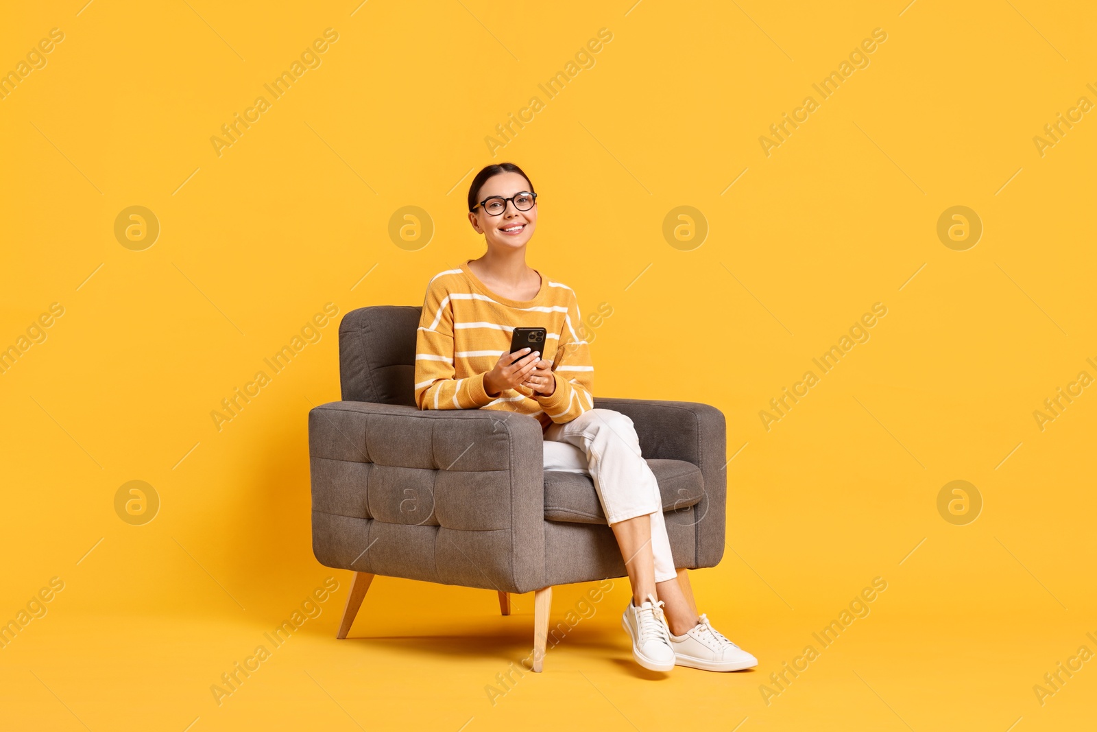 Photo of Smiling woman with smartphone sitting on armchair against yellow background