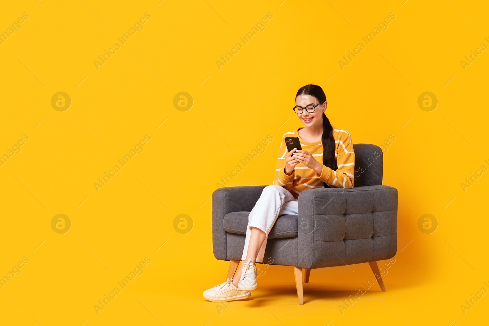 Photo of Smiling woman with smartphone sitting on armchair against yellow background. Space for text