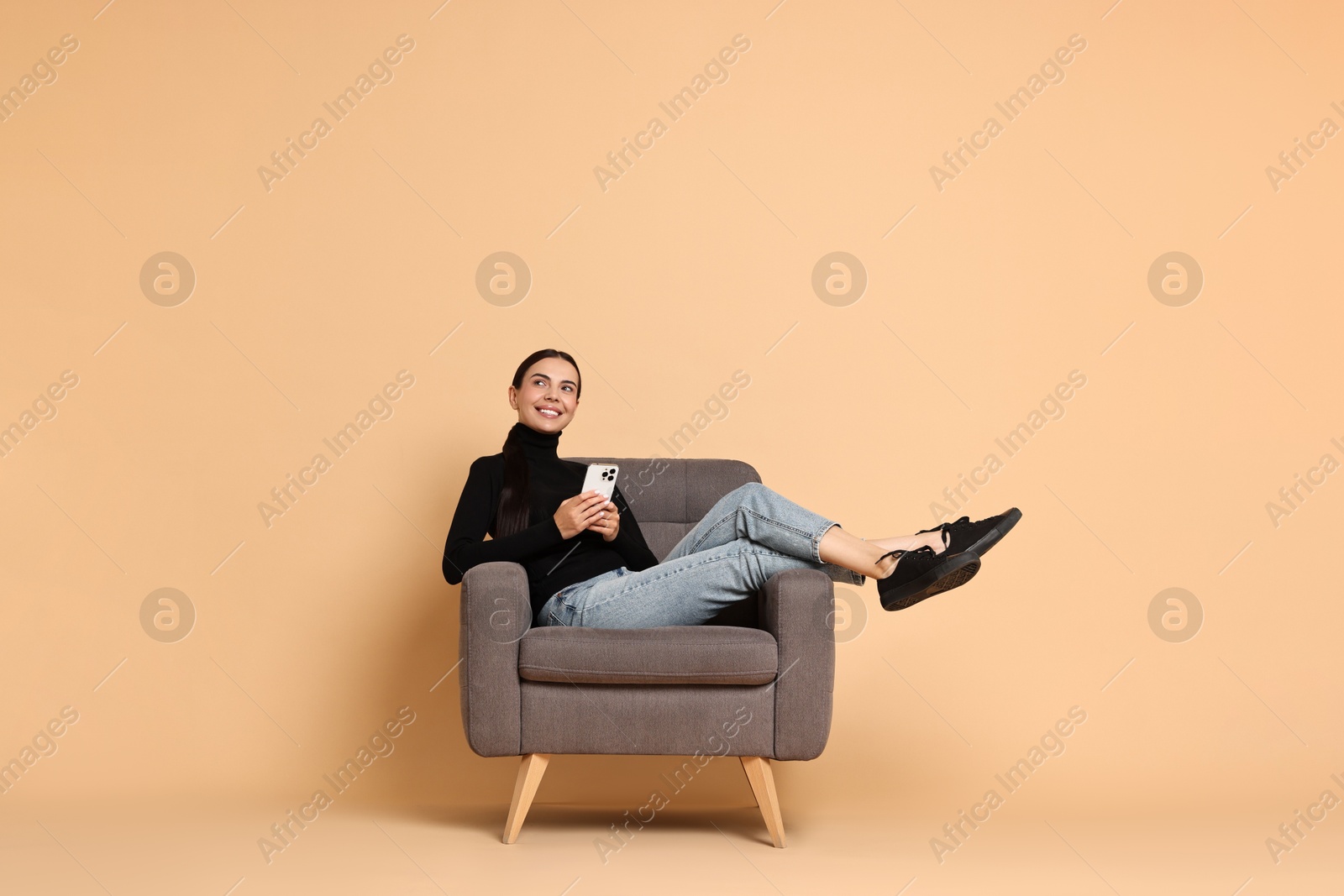 Photo of Smiling woman with smartphone sitting on armchair against beige background