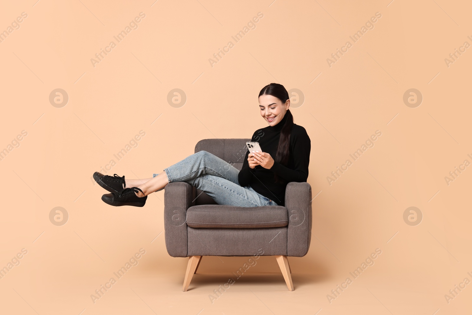 Photo of Smiling woman with smartphone sitting on armchair against beige background