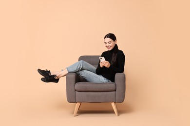 Smiling woman with smartphone sitting on armchair against beige background