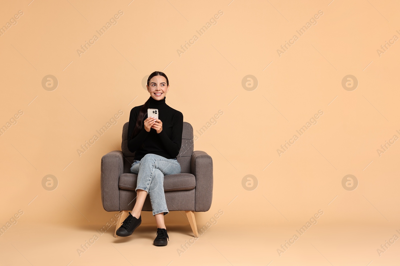 Photo of Smiling woman with smartphone sitting on armchair against beige background. Space for text