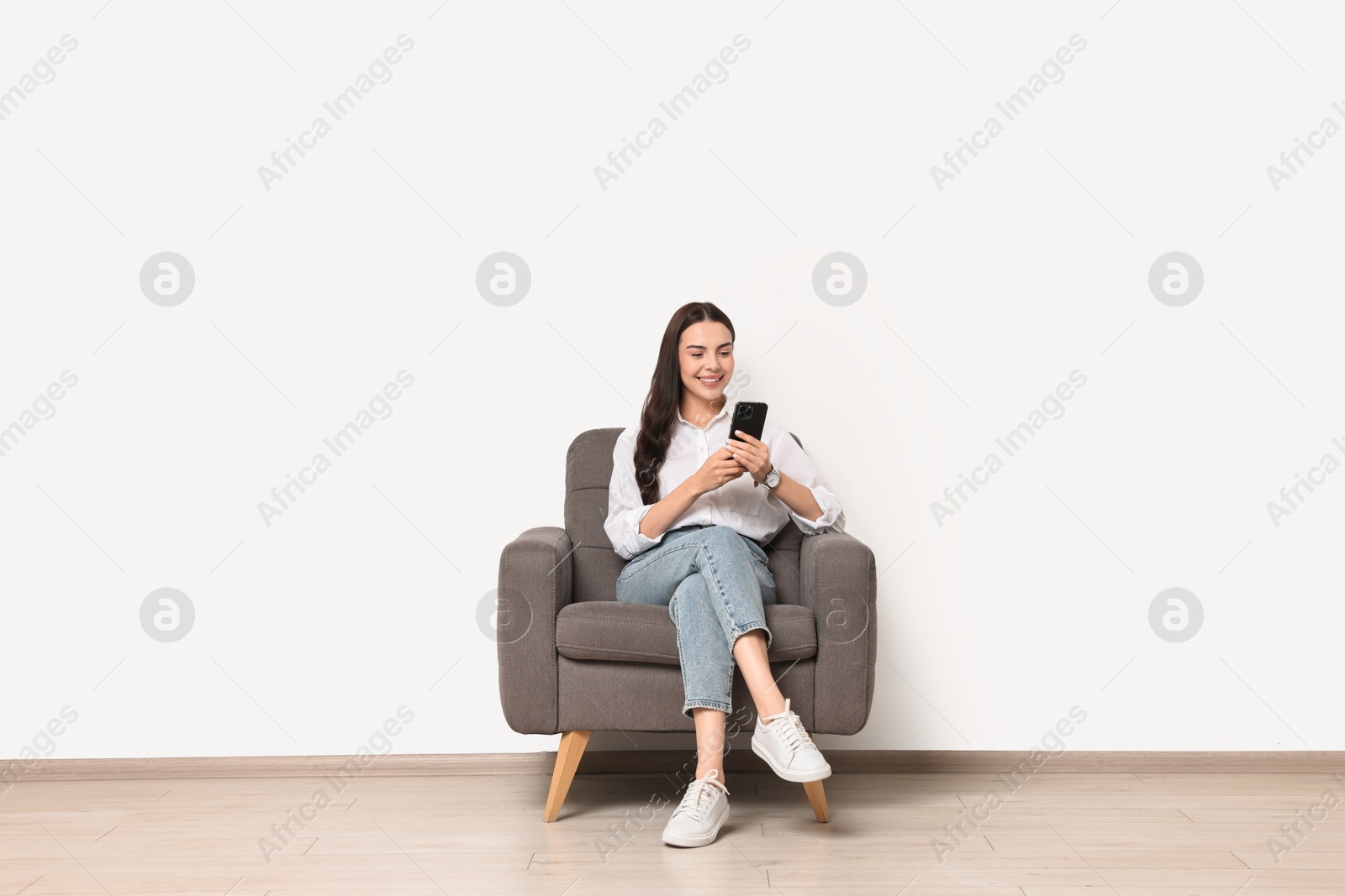 Photo of Smiling woman with smartphone sitting on armchair against white wall