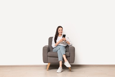 Photo of Smiling woman with smartphone sitting on armchair against white wall