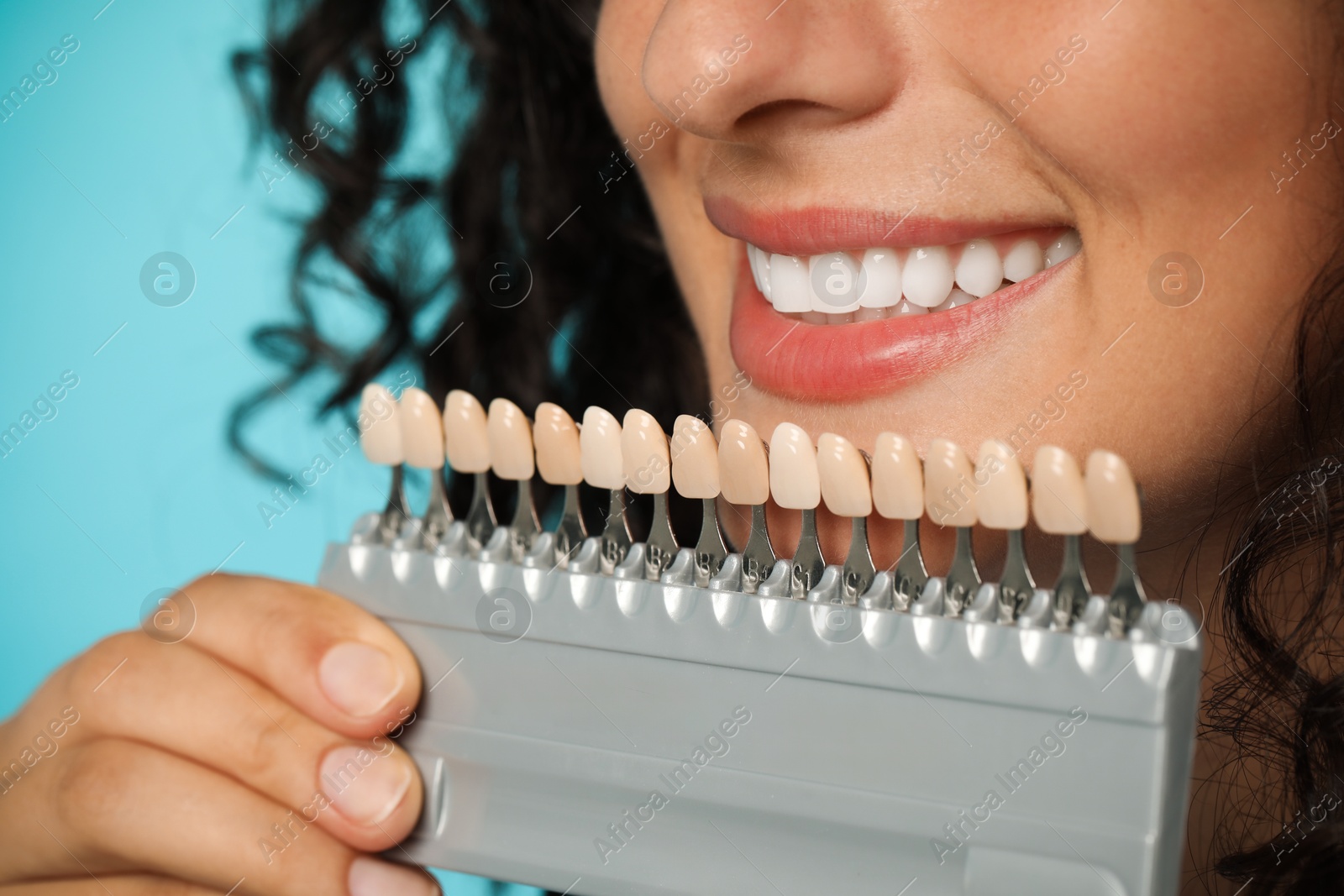 Photo of Smiling woman with teeth color samples on light blue background, closeup. Dental veneers