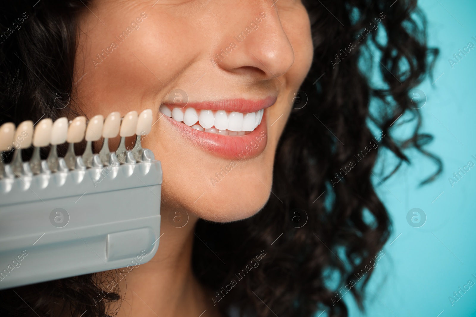 Photo of Smiling woman with teeth color samples on light blue background, closeup. Dental veneers