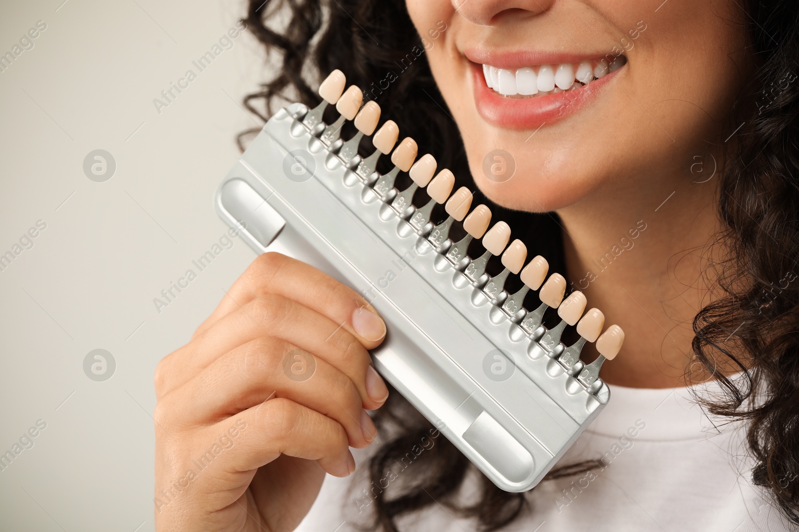 Photo of Smiling woman with teeth color samples on light grey background, closeup. Dental veneers