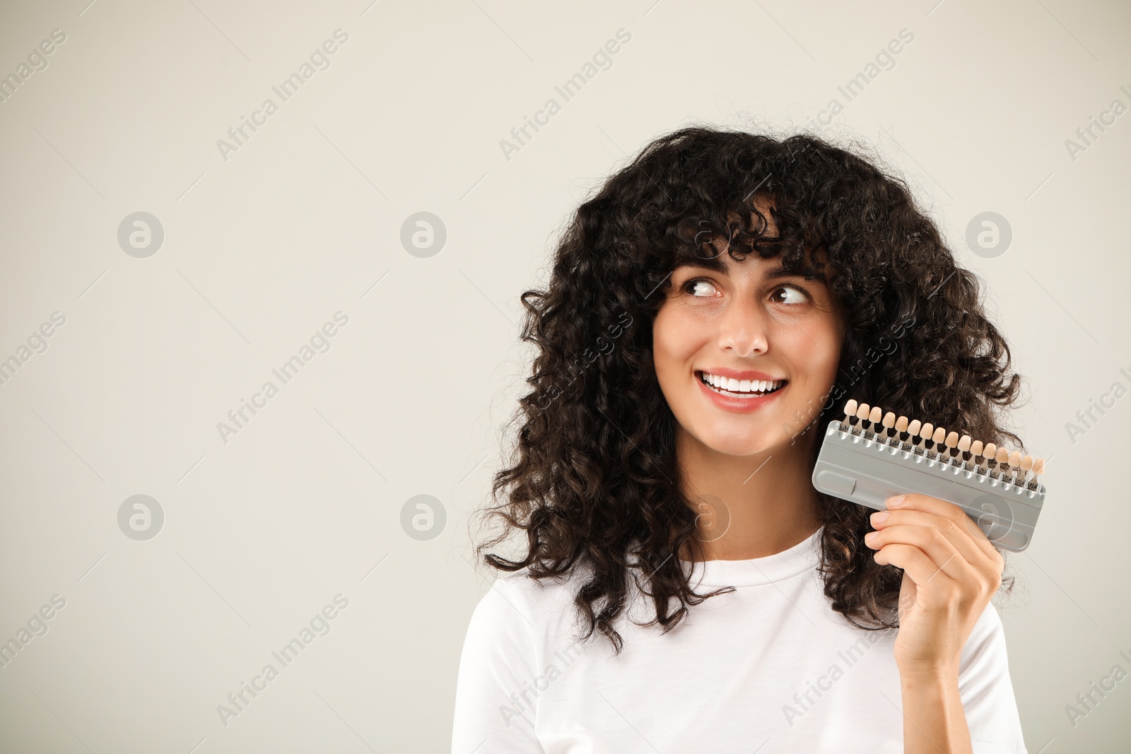 Photo of Happy young woman with teeth color samples on light grey background, space for text. Dental veneers