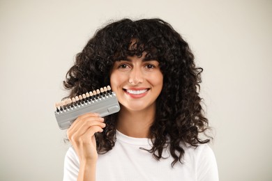 Happy young woman with teeth color samples on light grey background. Dental veneers