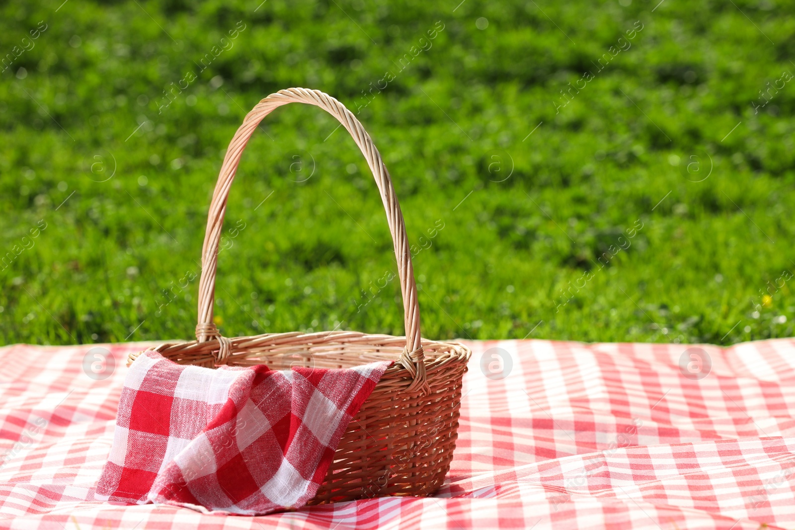 Photo of One picnic wicker basket with checkered napkin and blanket on green grass. Space for text