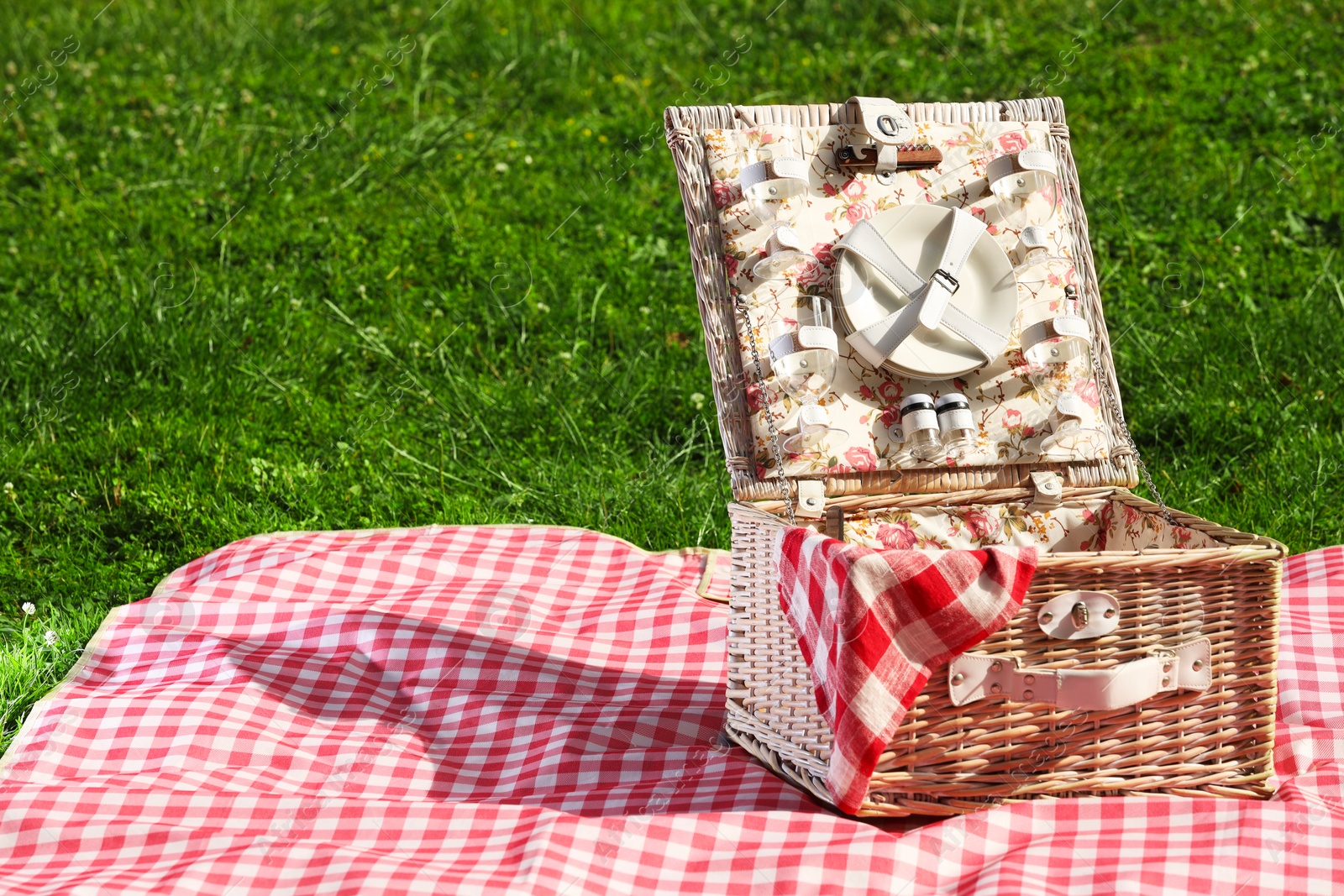 Photo of One picnic wicker basket with checkered napkin, tableware and blanket on green grass. Space for text