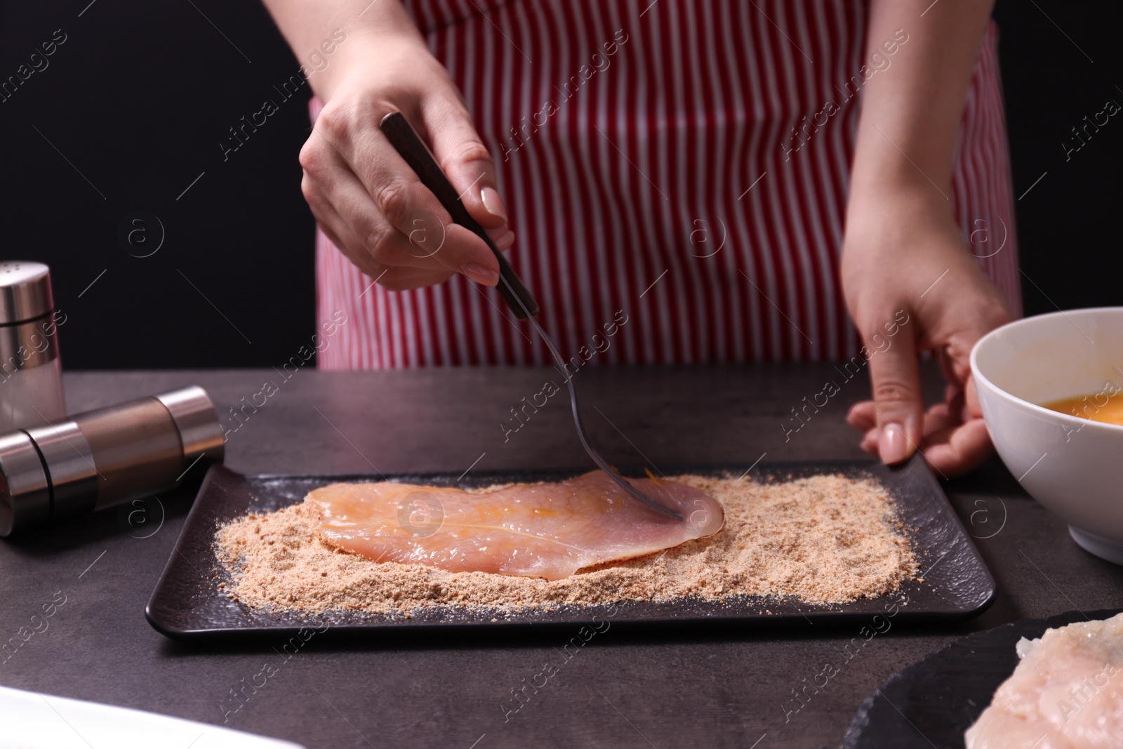 Photo of Making schnitzel. Woman coating slice of meat with bread crumbs at dark table, closeup