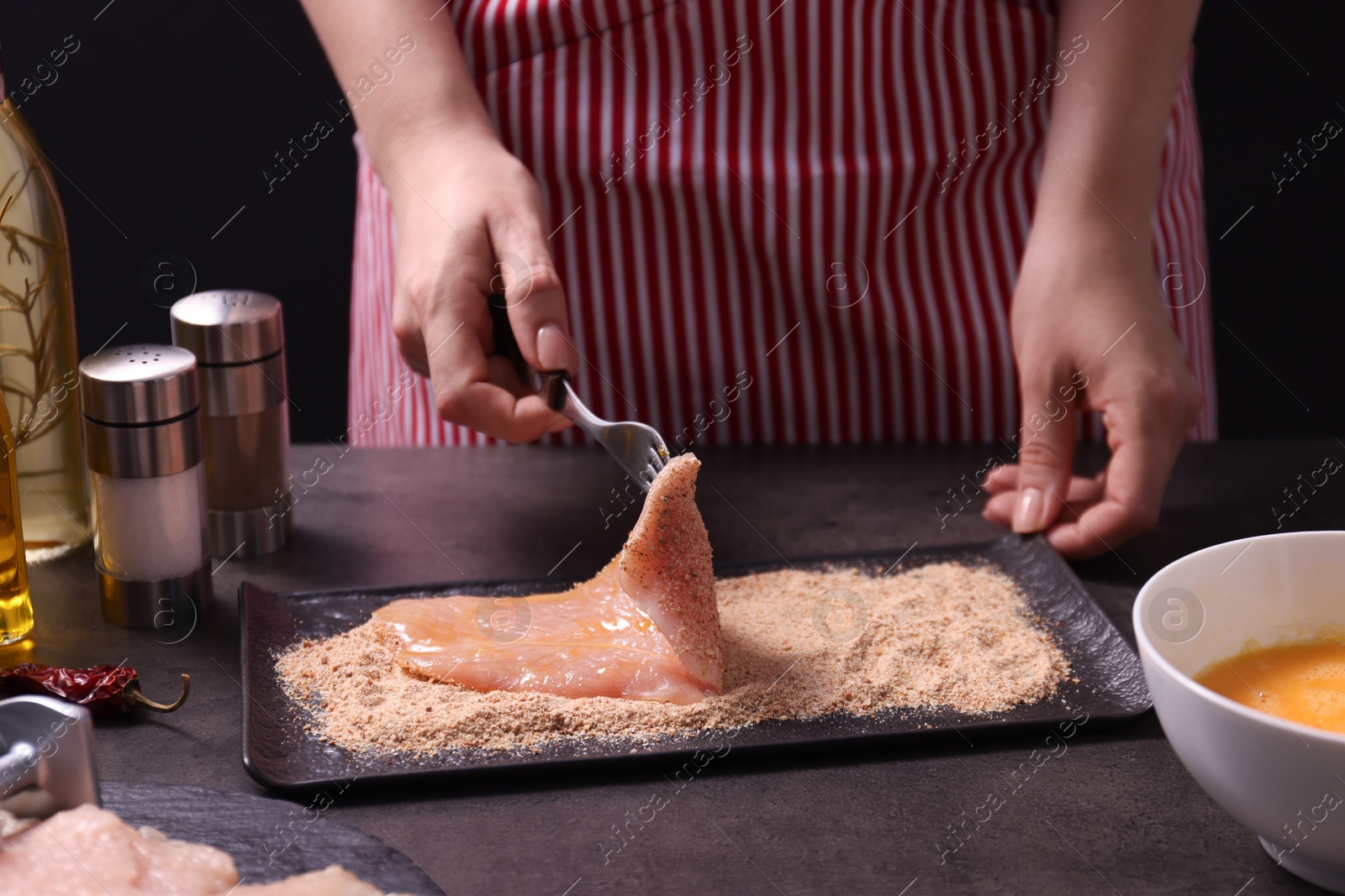 Photo of Making schnitzel. Woman coating slice of meat with bread crumbs at dark table, closeup