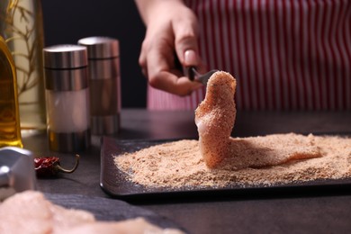 Making schnitzel. Woman coating slice of meat with bread crumbs at dark table, closeup