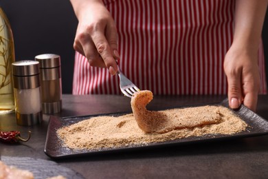 Making schnitzel. Woman coating slice of meat with bread crumbs at dark table, closeup