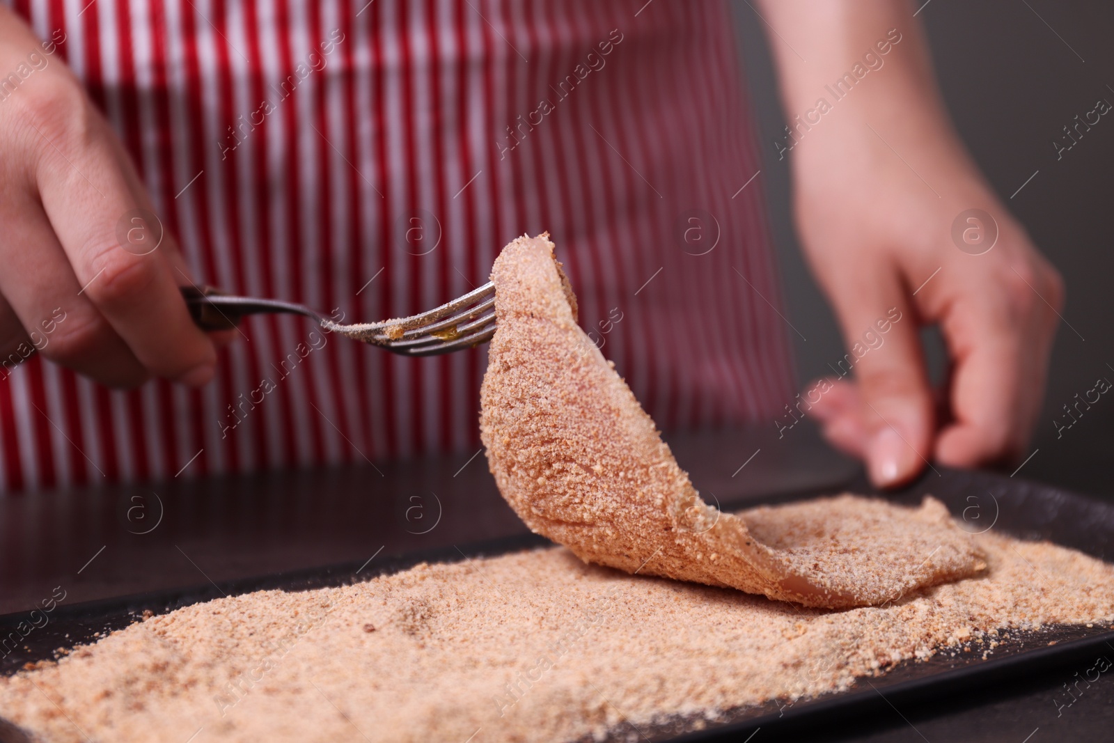 Photo of Making schnitzel. Woman coating slice of meat with bread crumbs at dark table, closeup