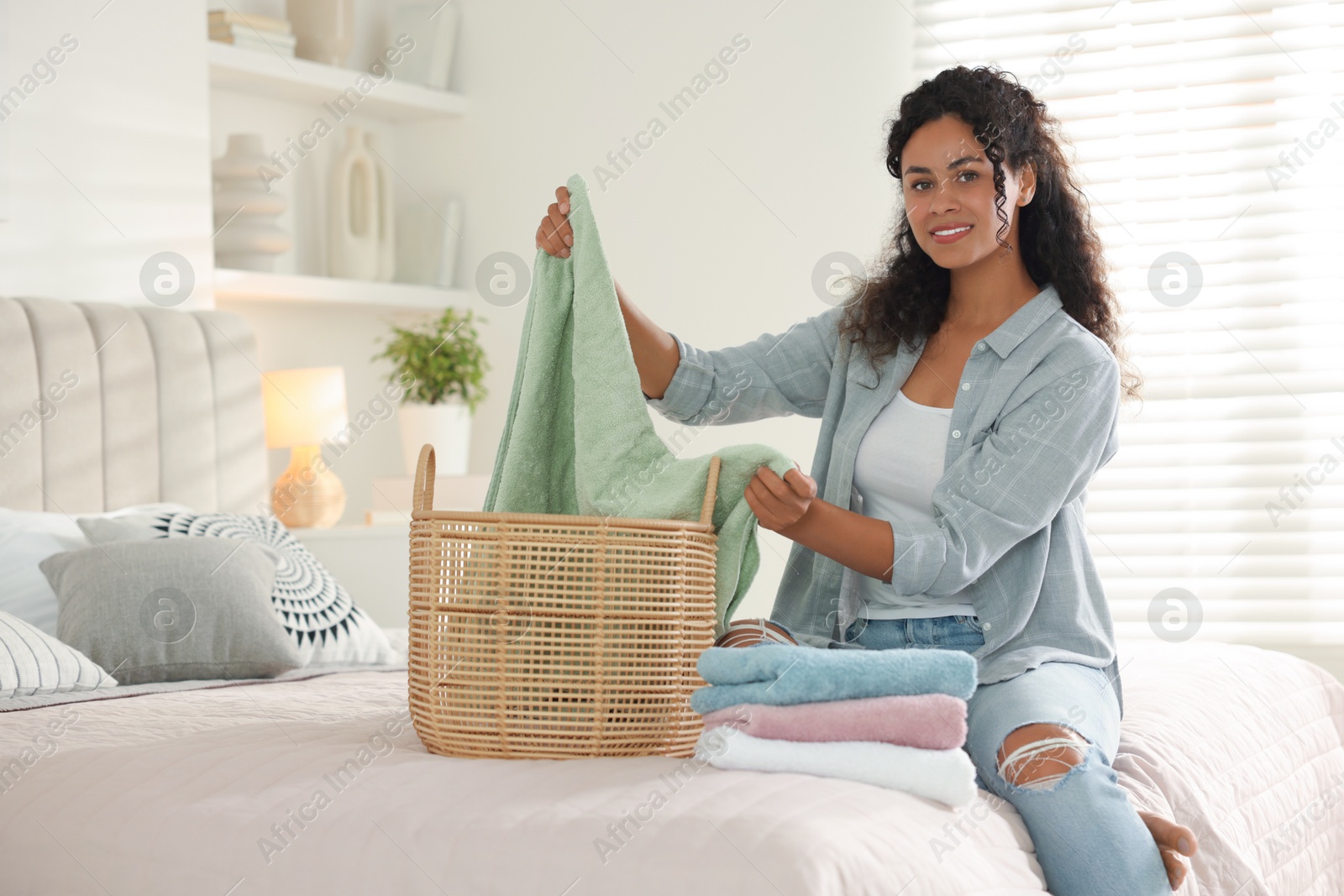 Photo of Happy woman with basket full of laundry on bed at home