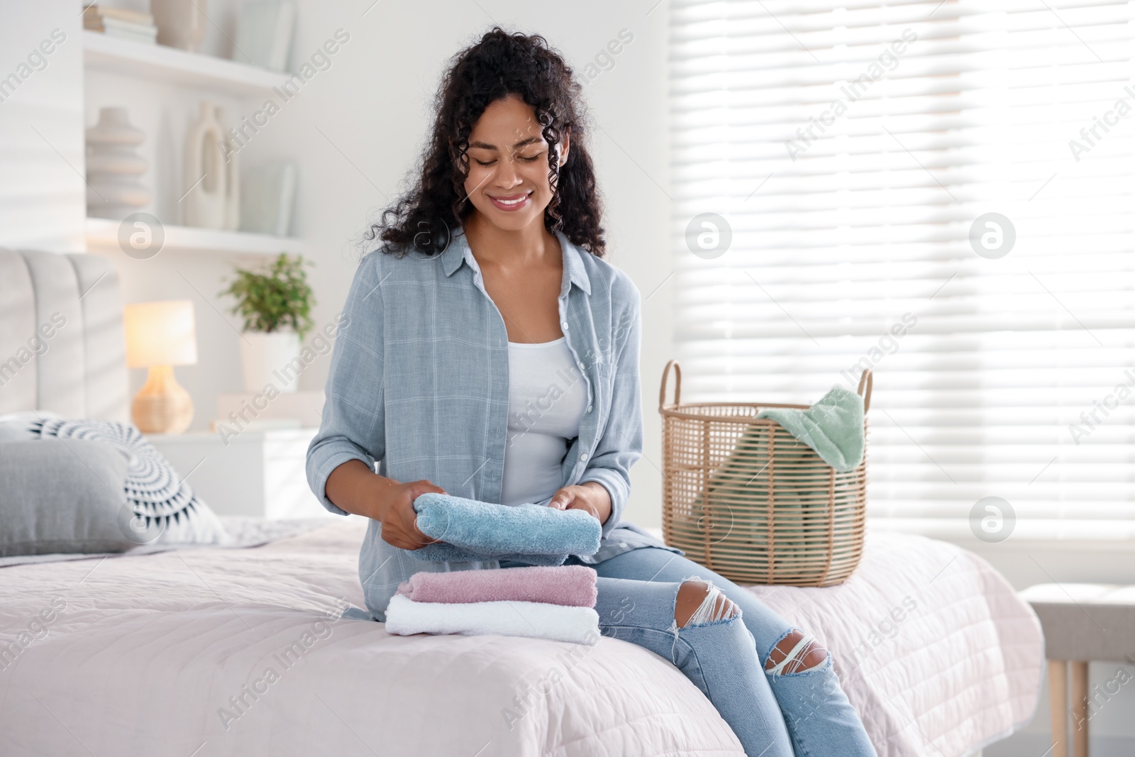 Photo of Happy woman with laundry near basket on bed at home