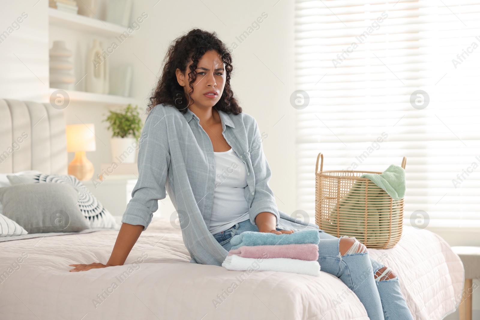 Photo of Displeased woman with laundry near basket on bed at home