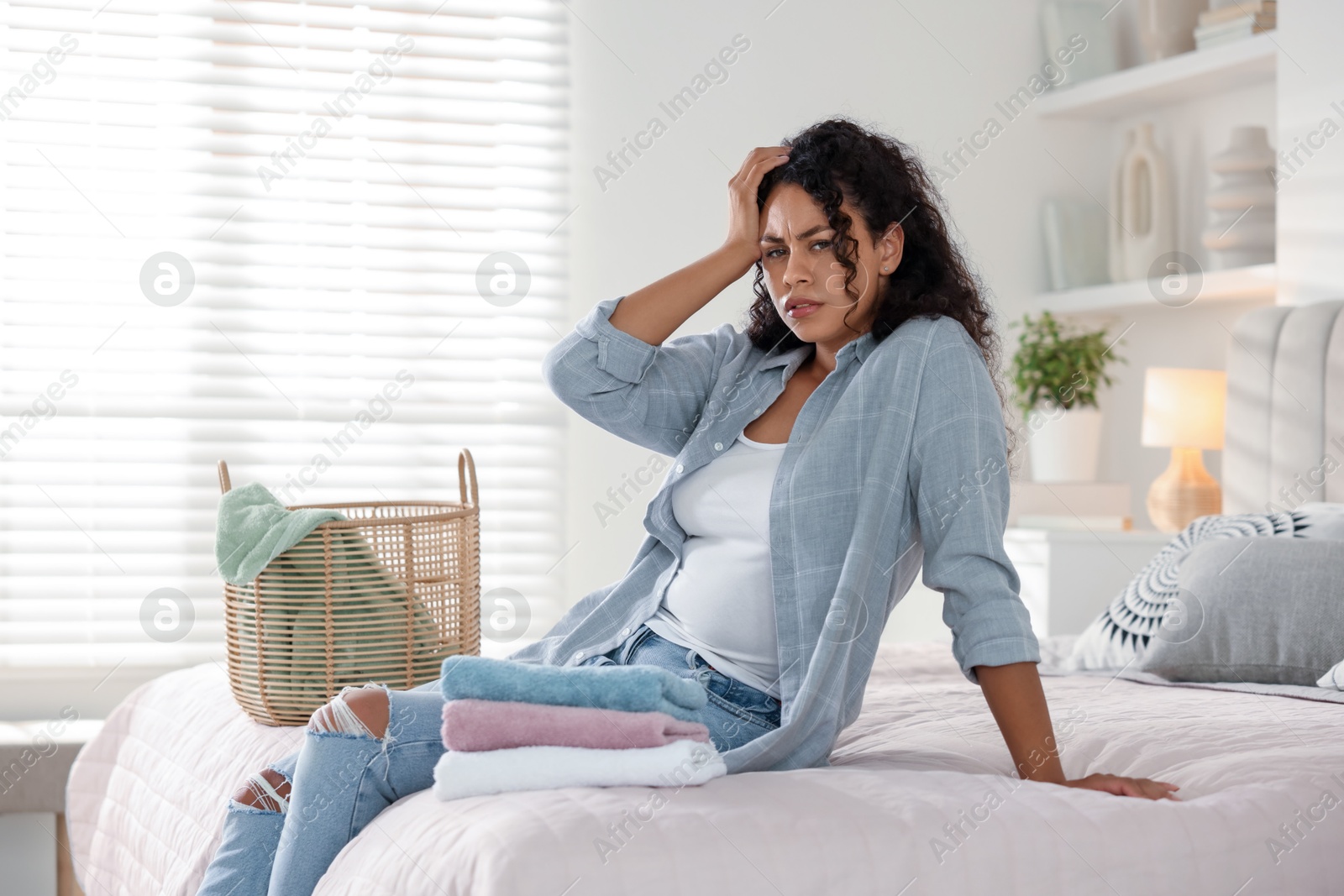 Photo of Displeased woman with laundry near basket on bed at home