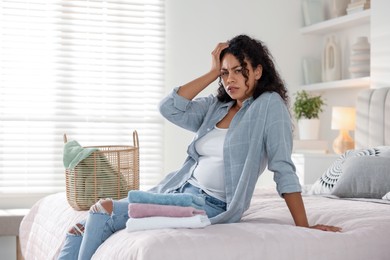 Displeased woman with laundry near basket on bed at home