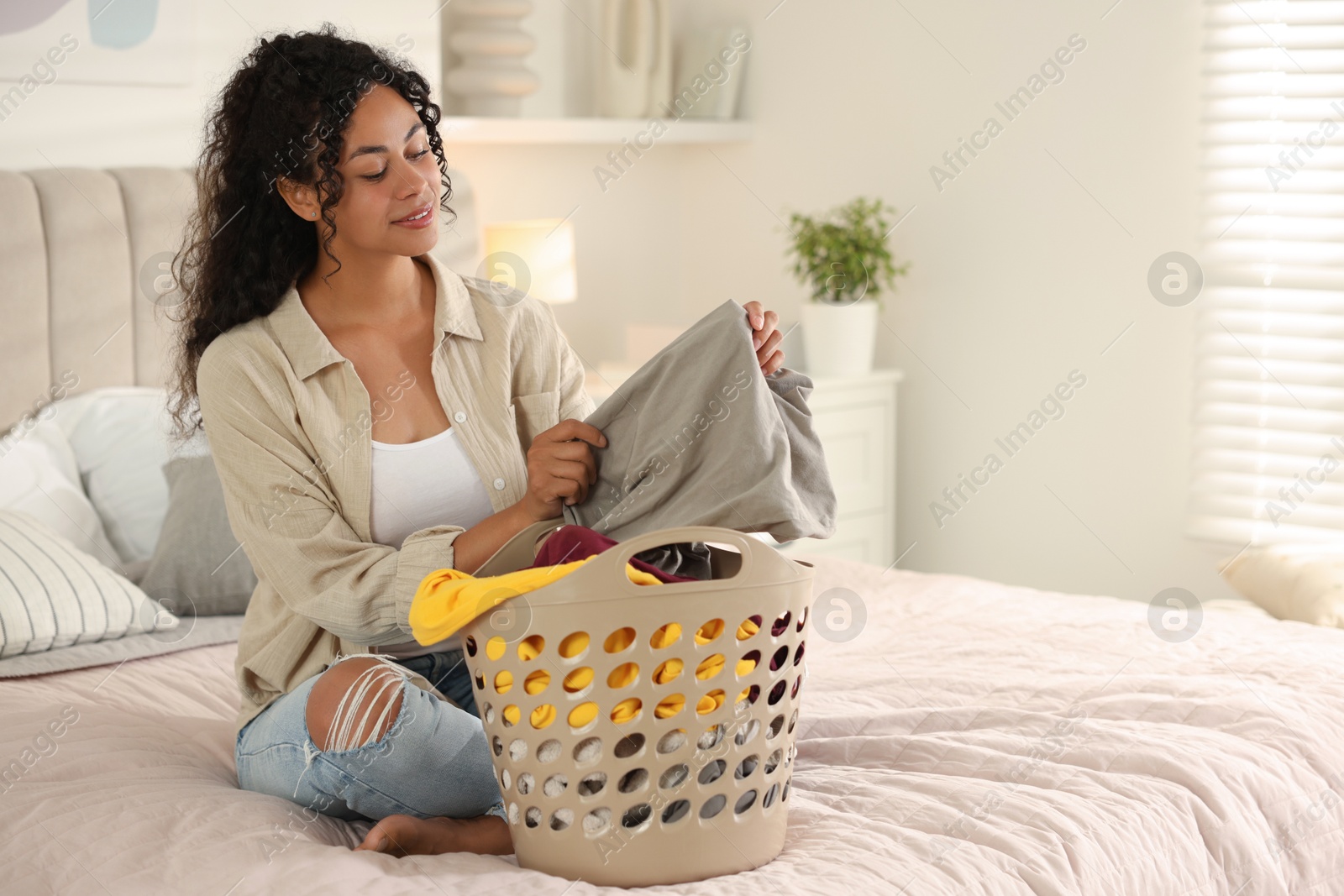 Photo of Happy woman with basket full of laundry on bed at home, space for text