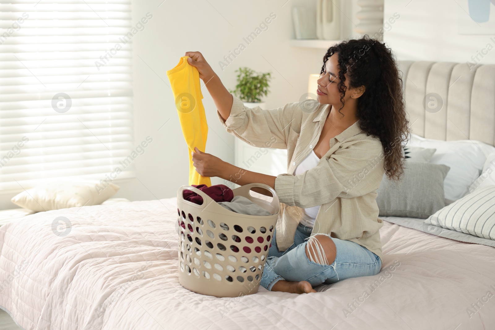 Photo of Happy woman with basket full of laundry on bed at home, space for text