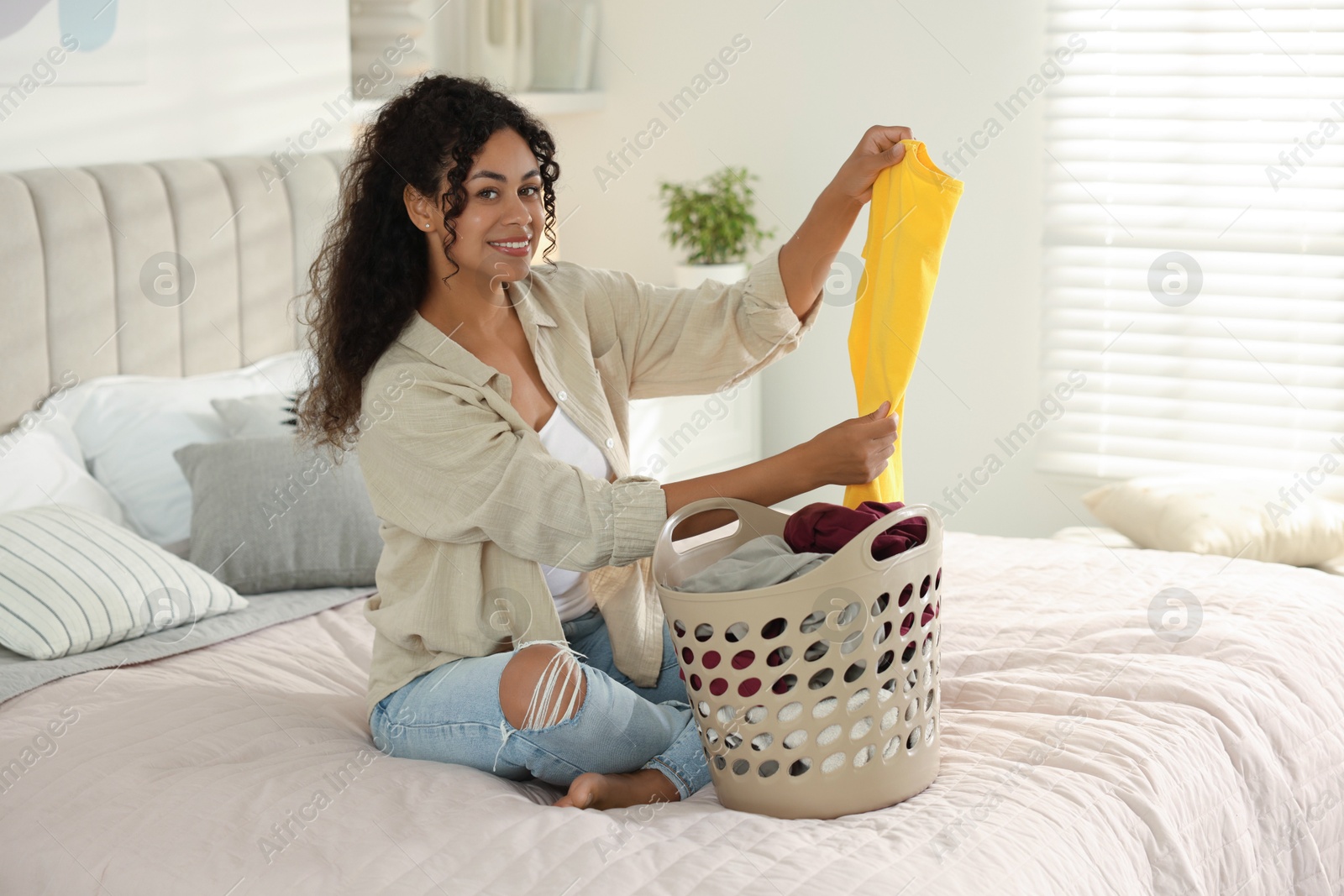 Photo of Happy woman with basket full of laundry on bed at home